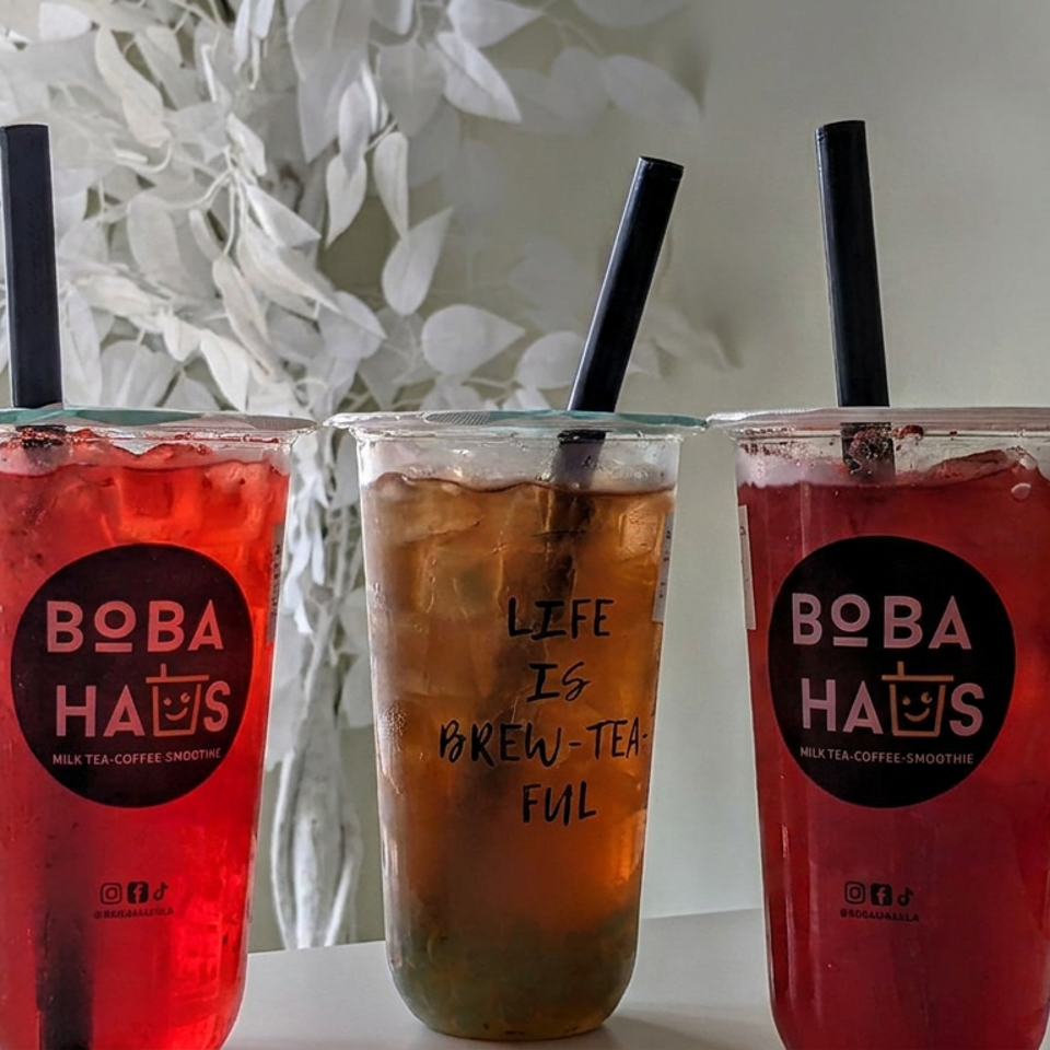 Three iced Boba Haus drinks in sealed cups, including two bright red beverages and one amber tea, displayed on a table.
